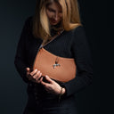 Femme regardant un sac bandoulière en cuir marron porté à l’épaule sur fond sombre.