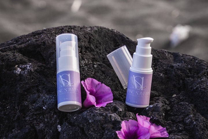 Produits Musc Soyeux en flacons pompe présentés en extérieur sur rochers avec fleurs violettes.